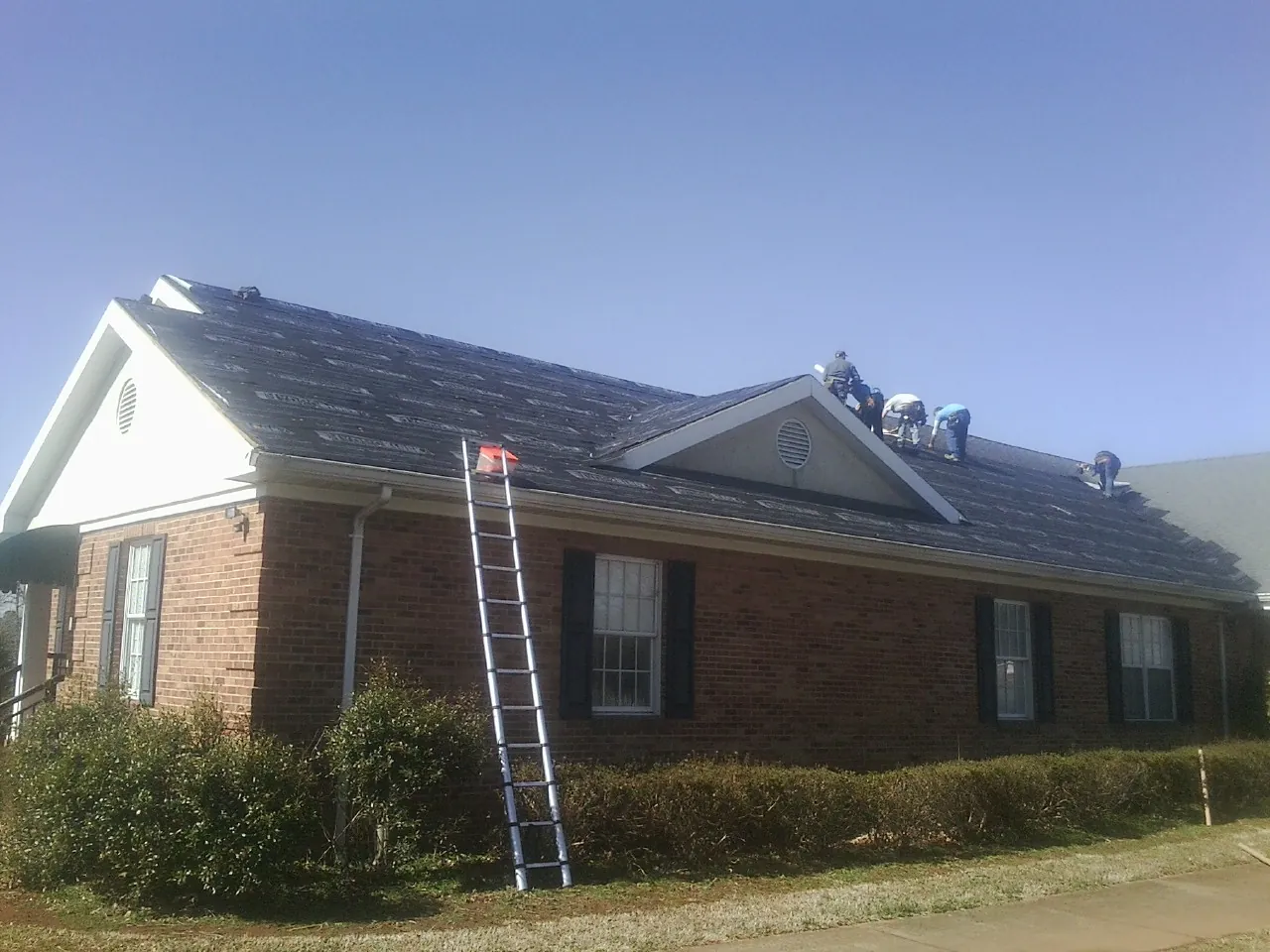 Crew tearing shingles off a brick ranch roof with a ladder set — Metal Roof Repair work by our crew in Erin